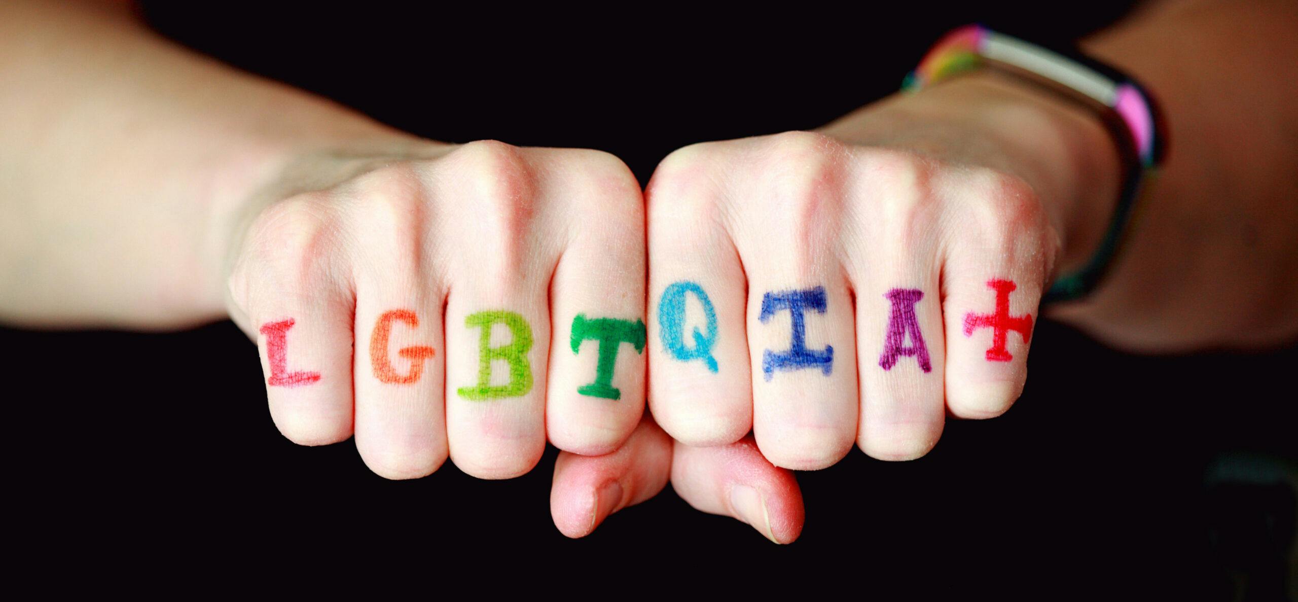 Two hands with the letters L G B T Q I A + written in rainbow colours on the knuckles