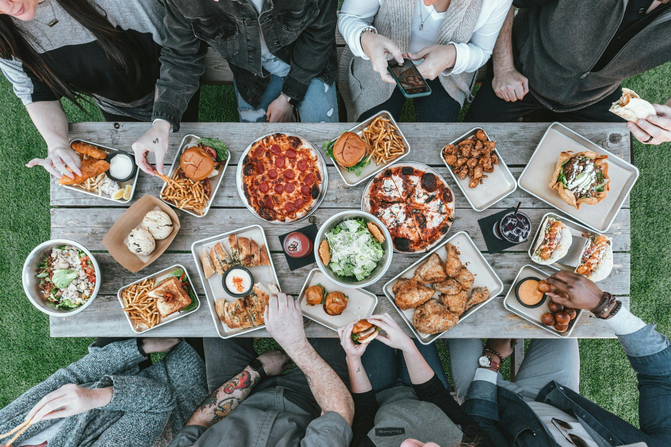 An aerial shot of eight people sat around a table eating pizza, fried chicken, and burgers.