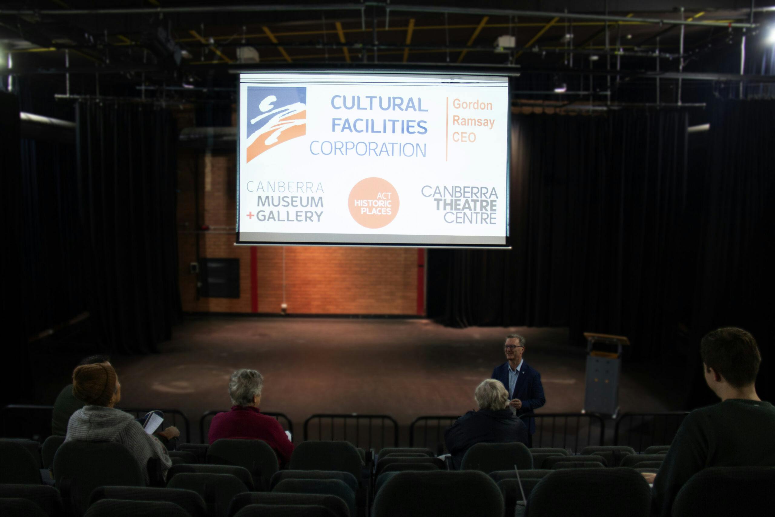 A small theatre as photographed from the back row - several people in the seats watching a man deliver a presentation.