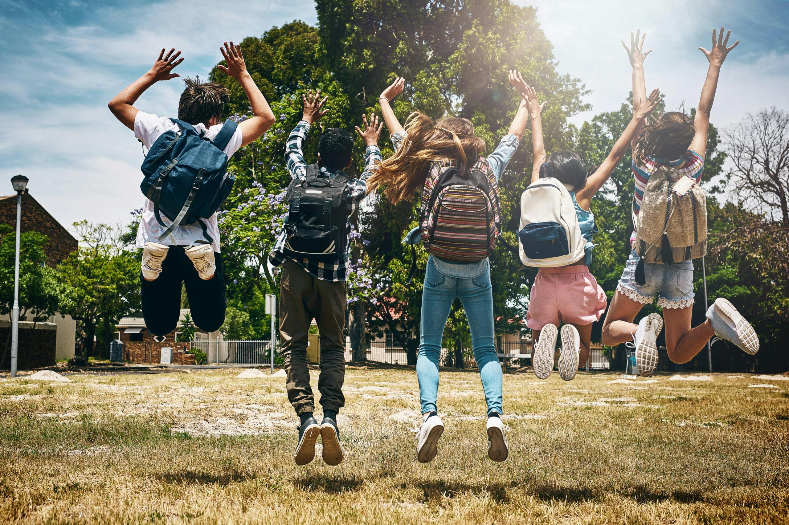 Five teens with school bags on, jumping in joy because it is now school holidays.
