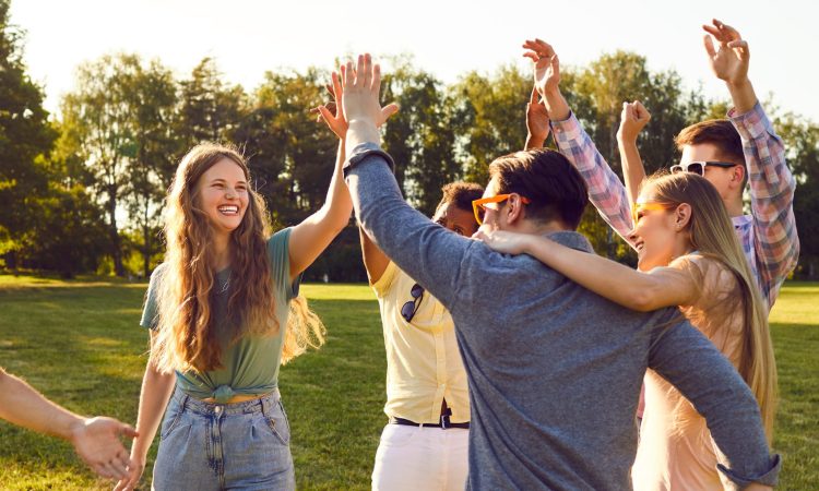 Bunch of happy young friends all together having fun on warm sunny day in summer park. Diverse group of cheerful joyful positive people standing on green lawn, smiling and giving each other high five