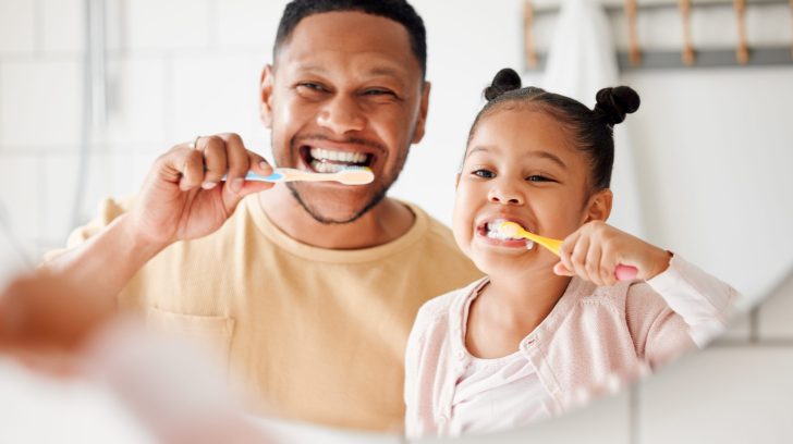 Child, dad and brushing teeth in a family home bathroom for dental health and wellness in a mirror. Face of african man and girl kid learning to clean mouth with toothbrush and smile for oral hygiene